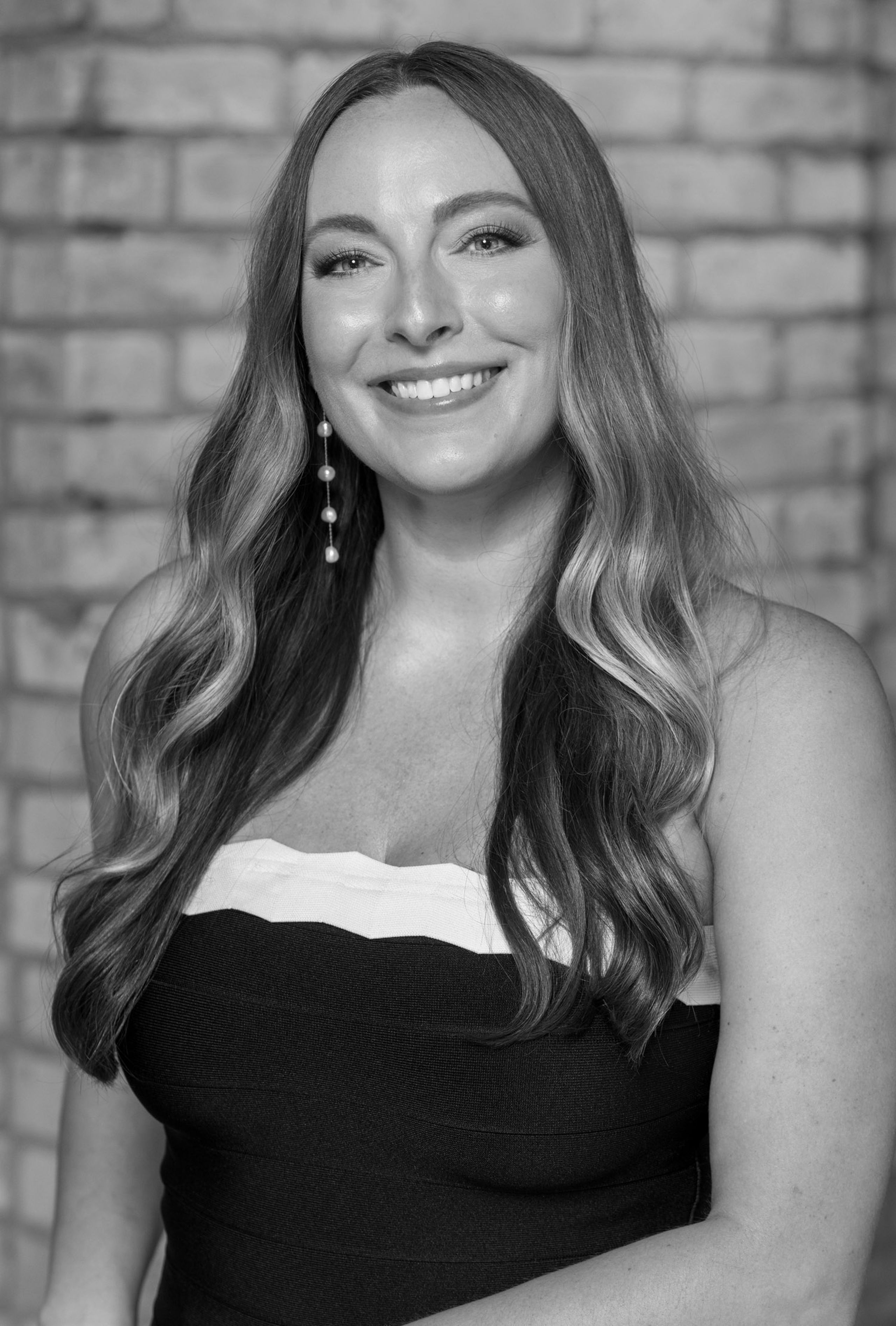 Black and white professional portrait of Jessi Pallan, Creative Director of Iron Diamond Media, wearing a strapless dress with long, wavy hair and pearl earrings, smiling in front of a brick wall.